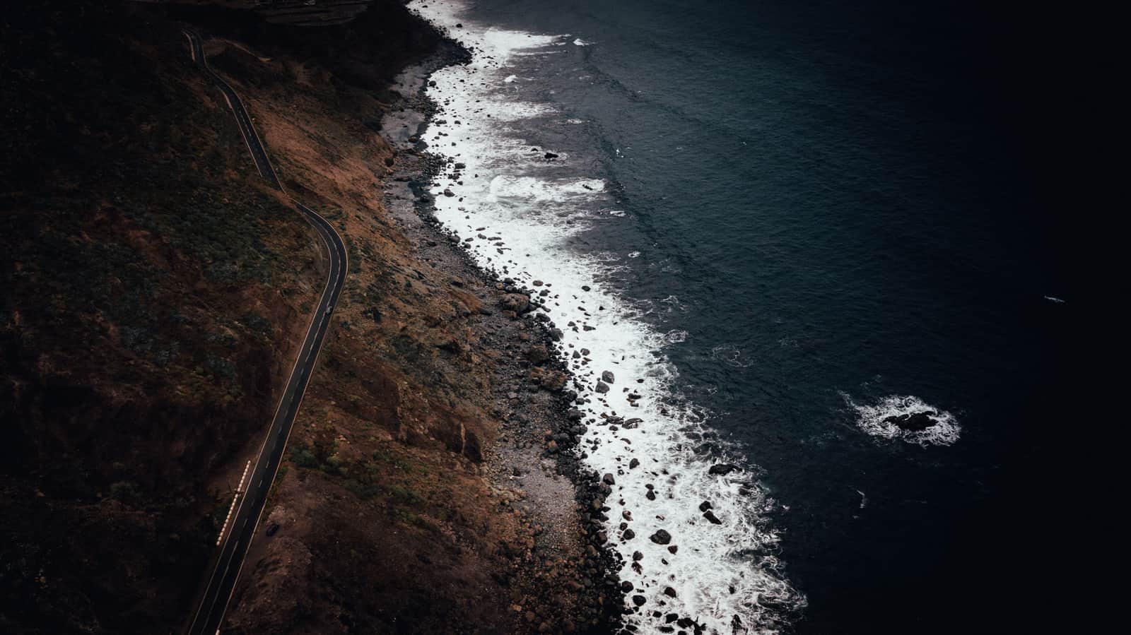 A road threading along a dark volcanic coastline seen from above