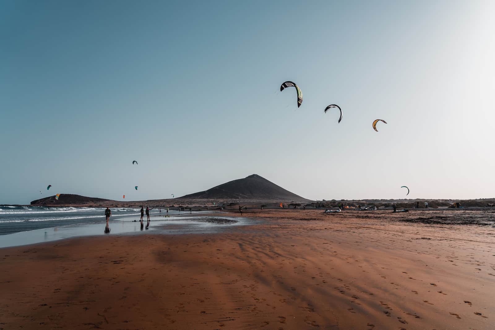 Kitesurfers on the wide flat beach below a volcanic cone